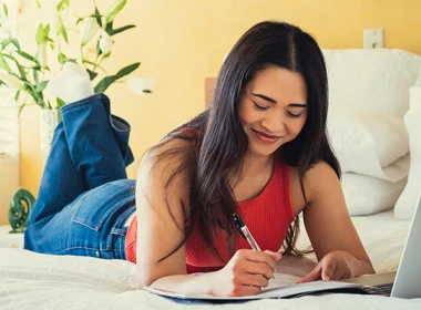 A woman is lying on a bed with her laptop and a notepad and pen