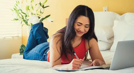 A woman is lying on a bed with her laptop and a notepad and pen