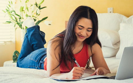 A woman is lying on a bed with her laptop and a notepad and pen