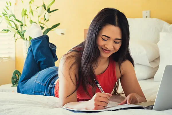 A woman is lying on a bed with her laptop and a notepad and pen