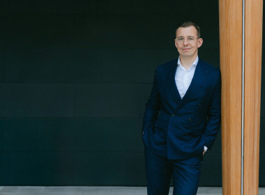 Alex in a navy suit and glasses leaning against a wooden pillar in a modern indoor setting.