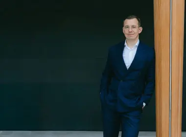 Alex in a navy suit and glasses leaning against a wooden pillar in a modern indoor setting.