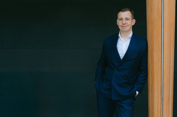 Alex in a navy suit and glasses leaning against a wooden pillar in a modern indoor setting.