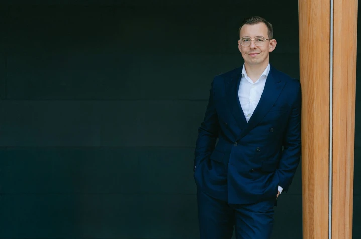 Alex in a navy suit and glasses leaning against a wooden pillar in a modern indoor setting.