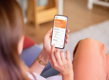 Over-the-shoulder view of a person sitting on a couch, holding a smartphone that displays Sorted's buffer builder app with an account balance and transaction details on the screen.