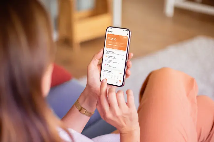Over-the-shoulder view of a person sitting on a couch, holding a smartphone that displays Sorted's buffer builder app with an account balance and transaction details on the screen.