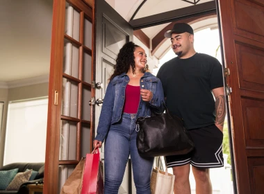 Couple look at each other while holding shopping bags and walking into a home. 