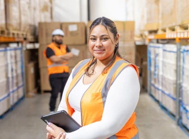 Woman smiling at the camera, wearing an orange high‑visibility vest, with a blurred factory background.