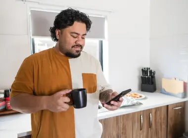 Person standing in a kitchen holding a black mug and checking a smartphone, with kitchen benches, cupboards, and food items visible in the background.
