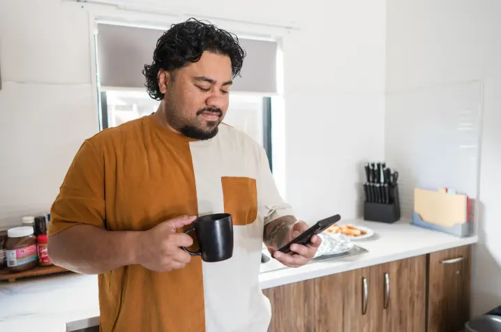 Person standing in a kitchen holding a black mug and checking a smartphone, with kitchen benches, cupboards, and food items visible in the background.