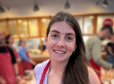 Gemma smiling and looking at the camera, wearing a red apron and holding a fresh pack of pasta