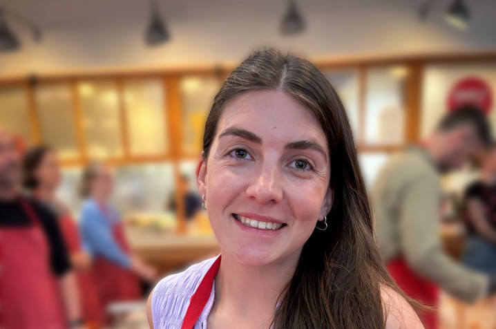 Gemma smiling and looking at the camera, wearing a red apron and holding a fresh pack of pasta