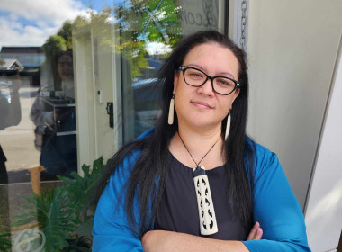 Indigo Phillips standing outside a building, wearing a blue jacket and pendant, representing a Māori woman sharing her homeownership journey with Sorted Kāinga Ora. 