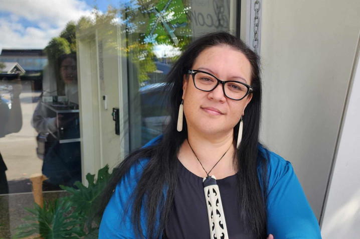 Indigo Phillips standing outside a building, wearing a blue jacket and pendant, representing a Māori woman sharing her homeownership journey with Sorted Kāinga Ora. 