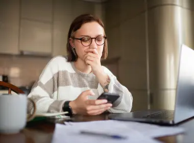 Woman seated at a table, holding a smartphone and looking at it with worried expression. 