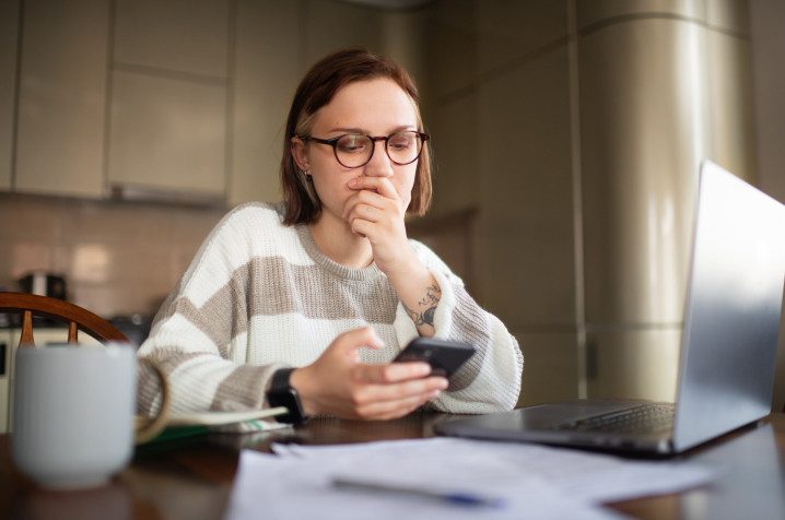 Woman seated at a table, holding a smartphone and looking at it with worried expression. 