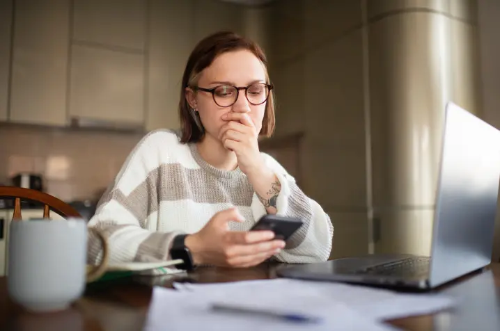 Woman seated at a table, holding a smartphone and looking at it with worried expression. 
