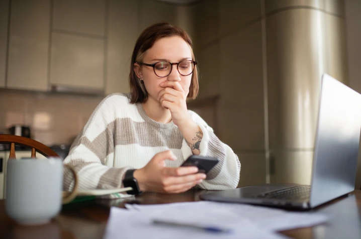 Woman seated at a table, holding a smartphone and looking at it with worried expression. 