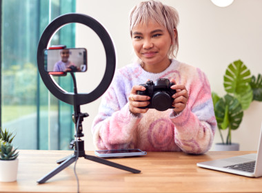 Young woman holding a professional camera, surrounded by a laptop, studio lighting, and a phone recording, capturing content for a side hustle.