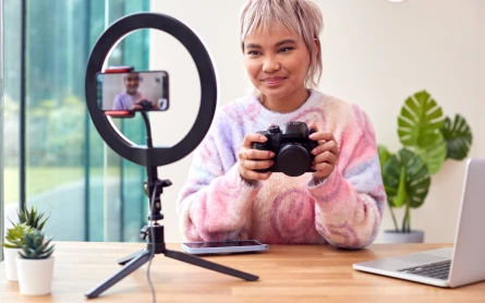 Young woman holding a professional camera, surrounded by a laptop, studio lighting, and a phone recording, capturing content for a side hustle.