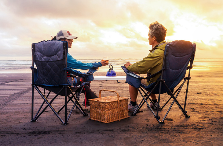 Two people sitting in folding chairs on a sandy beach at sunset, facing the ocean, with a picnic basket and a teapot on a small table between them.