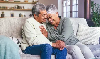 Two adults sitting close together on a couch indoors, holding hands with a kitchen and living room visible in the background