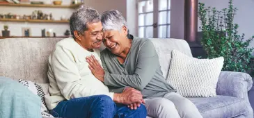 Two adults sitting close together on a couch indoors, holding hands with a kitchen and living room visible in the background